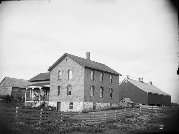 View from field of home and farm of Henry Hoff. Men and women stand on the porch of the large brick house. Beyond the house is a small log barn, dwarfed by the large frame barn behind it.