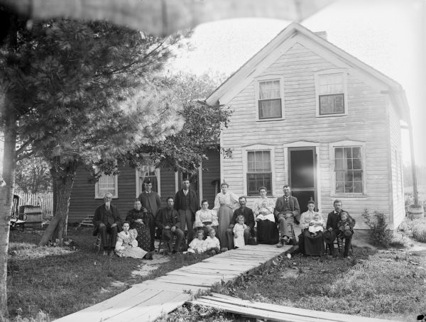 A family group of 19 people pose in front of their house. The photographer's umbrella can be seen at the top of the frame.