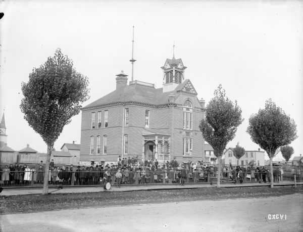 View across road towards the exterior of a brick elementary school in northern Wisconsin. A large group of children and adults pose from within the schoolyard fence. A number of boys are standing on the sidewalk in the foreground.