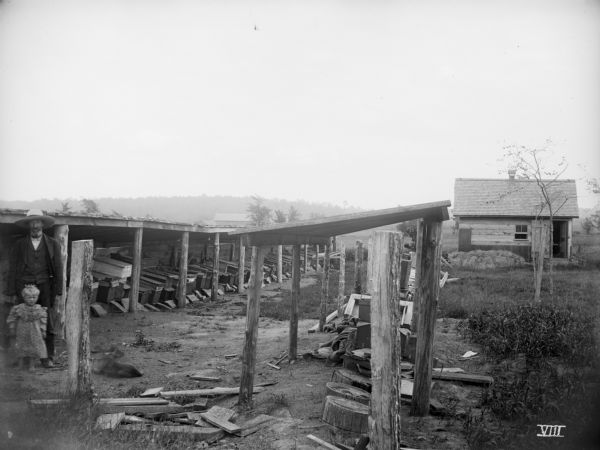 A man, child and dog pose between two long sheds. Underneath the sheds are rows of wooden boxes.