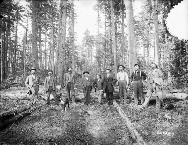 A group of well-dressed men and a dog pose in a forested area.