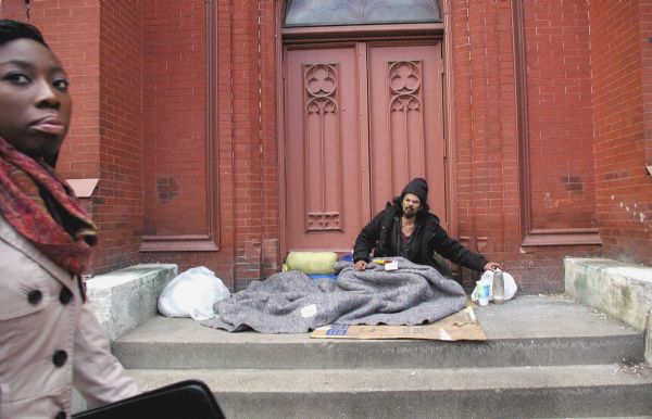 A homeless man is sitting on Saint Patrick's Catholic Church's steps as a woman on the sidewalk is walking past.