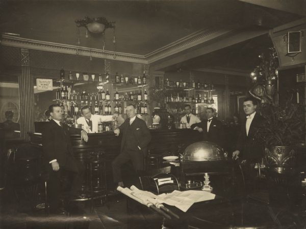 Six men standing in a dining room with a bar. Two of the men are bartenders, one of whom is pouring a drink. Three men dressed in suits are leaning against the bar, and one waiter is pushing a serving cart. Also visible in the foreground is a table that is piled with newspapers. Some of the men are identified on the back of the photograph. Left to Right, 1. Luigi, 2. John Blackmen (?), 3. Captain A. Client, 4. Pittom, 5. Dell (?), 6. ?.