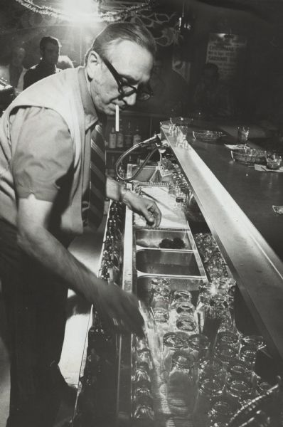 Bartender Albert Rosen cleaning glassware behind the bar. Rosen has a cigarette in his mouth as he cleans. Men can be seen in the background at the bar. This image was featured in the "Milwaukee Sentinal" with the following caption: "Glasses filled the sink of Sardino's South, as Jewish Albert Rosen tended bar so that John Volpe could be free to spend Christmas Eve at home with his family."