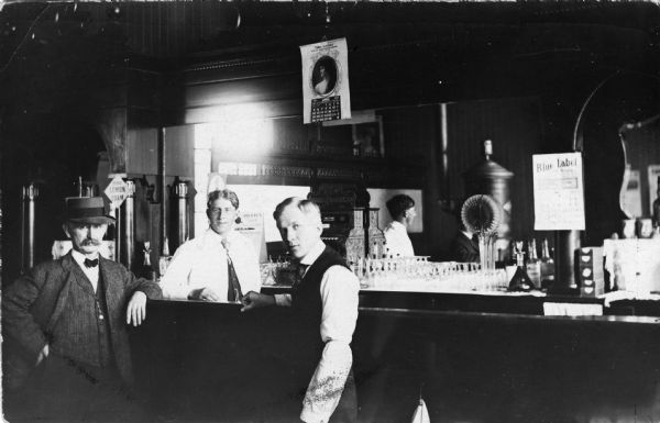 Two men standing at a bar with a bartender. The back of the bar is mirrored and reflects some of the room, with a wood stove seen in the reflection. A cash register and glassware are behind the bar along with a few glass bottles. Hanging over the bar is a calendar with an image of a women on the front. The calendar reads April 1908.