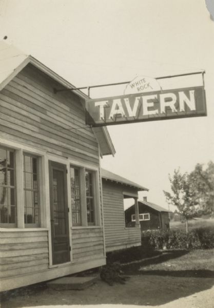 Exterior view of the White Rock Tavern. A large dog is laying in the shade of the building entrance. A large neon sign hangs from the front of the building with the name of the tavern. The caption on the back of the photograph reads, "The sign on brother tavern and a little of it before it was painted."