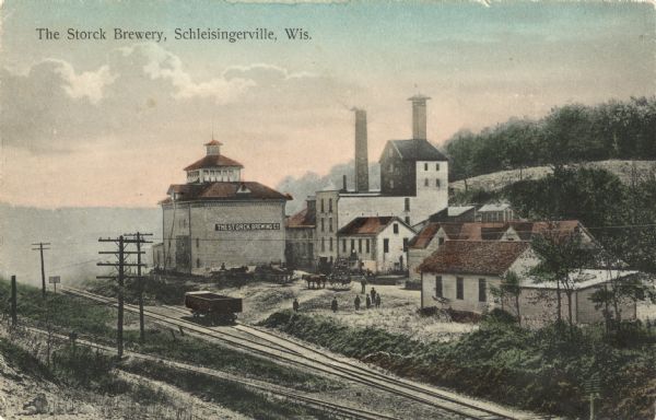 Colorized postcard view of the Storck Brewery in Schleisingerville (now Slinger). The brewery buildings are along a hillside, and railroad tracks are in the foreground. There is a sign on the side of one building that reads: "The Storck Brewing Co." Horses with carts and men can be seen among the brewery buildings. Caption reads: "The Storck Brewery, Schleisingerville, Wis."
