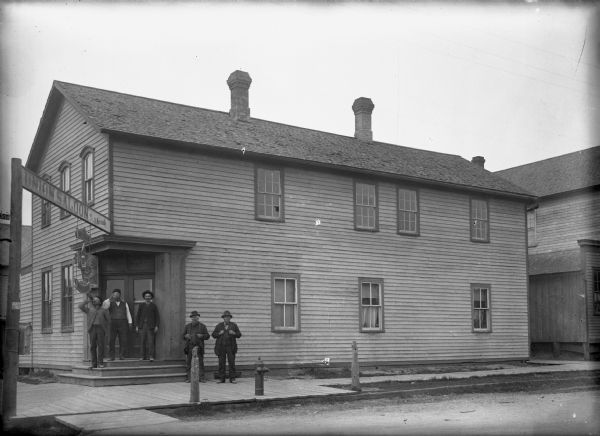 Exterior image of the Union Saloon. The saloon was owned by Joseph Krainik. Three men are standing on the entrance steps of the building and two men are standing on a plank sidewalk. Two of the men are drinking from bottles. There is a large sign the hangs over the sidewalk that reads "Union Saloon, by J. Krainik". The bar was on the corner, with an address of 401 N. 8th. The lower glass windows along the side of the building have interior shutters, and two of the upper windows have broken panes of glass.