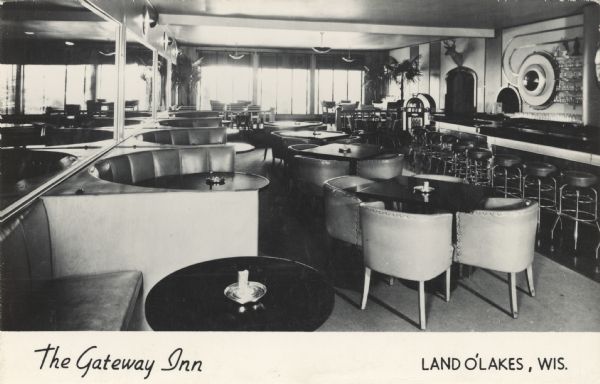 Postcard of an empty bar and lounge. In the foreground are a number of circular seating areas of booths along a mirrored wall. The bar runs along the right hand side. There is a jukebox near an arched doorway above which is displayed a deer head mount. Caption reads: "The Gateway Inn, Land O' Lakes, Wis."