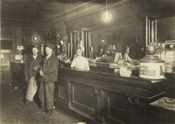 Interior of a bar in Northern Wisconsin. Two bar patrons are pictured standing in front of the bar, with a bartender posing behind them. The back of the carved wood bar is mirrored, and there is a large container sitting on the counter printed with the words: "Lash's Orangeade."