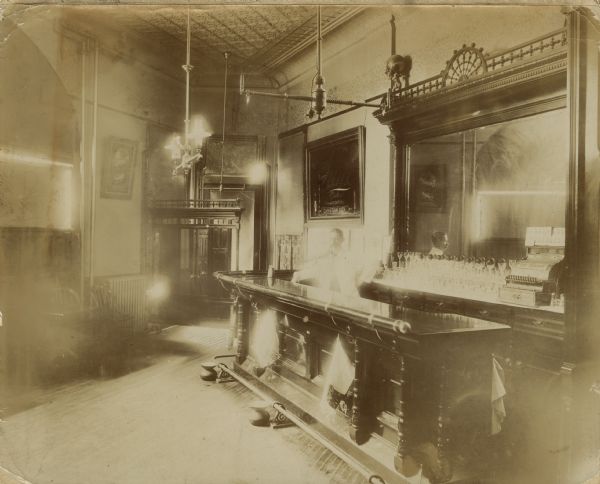 Man standing behind the bar at "The Schlitz Saloon" located on the corner of Doty and King Streets. The bar is backed with a large mirror and is lined with glasses and a cash register. Spittoons are on the floor along the foot rail in front of the bar.