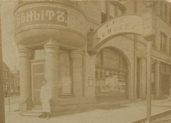 The Schlitz Saloon on the corner of Doty and King Streets. The saloon was owned by Fred C. Harbort from 1900-1907. There is a man in an apron standing at the door of the saloon. Above him on the right is a large arched sign that says "The Schlitz" which hangs over the sidewalk and is supported by a post coming up from the street.