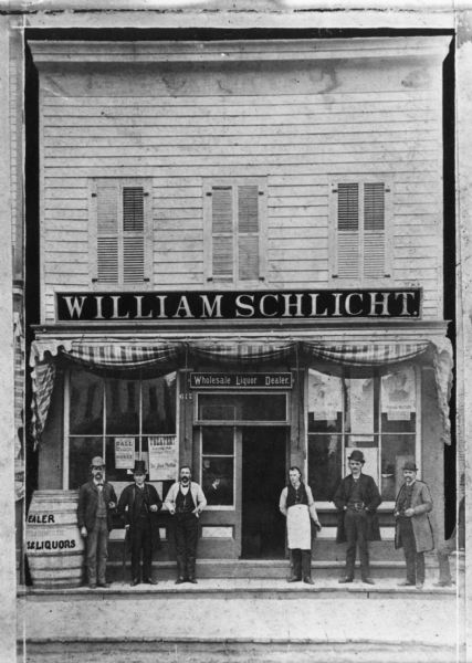 The storefront of the William Schlicht Wholesale Liquor Dealer. The building also was a saloon and features shuttered windows, advertising posters in the lower windows and a striped awning. Six men are standing on the planked sidewalk. Three of the men are wearing bowler hats, one is wearing a top hat and one man is wearing an apron.
