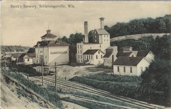 Postcard of the Stork Brewery facilities. The image includes the brewery buildings along a hillside and railroad tracks in the foreground. There is a sign on the side of one building that reads: "The Storck Brewing Co." Caption reads: "Stork's Brewery, Schleisingerville, Wis."