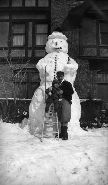 Two children posing in front of a large snowman in front of a brick building. The smaller child is sitting on a ladder supported by the older boy. The snowman is decorated with a hat, cane, pipe and a Schlitz Beer bottle.