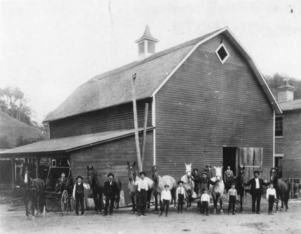 Group portrait of men and boys pictured with horses outside a barn. The stables served the Potosi Brewing Company. On the far left a man is seated in a carriage. There is a two-story brick building on the right, and a steep hill in the background on the left.