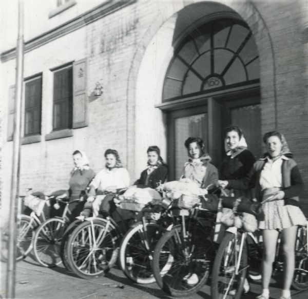Six girls with their bicycles standing in front of Neighborhood House at 768 W. Washington Avenue, ready to depart on the first bike hosteling trip sponsored by the settlement house. Trip participants were Donna Jean McCann, Vincella Raimond, Vincenza Raimond, Mary Pecoraro, Teresa Mandarino, Mary Baldarato, and group leader Martha Nelson (probably not pictured here). Cyclists pedaled to Pine Bluff and Cross Plains before returning to Madison on a 55-mile roundtrip.
