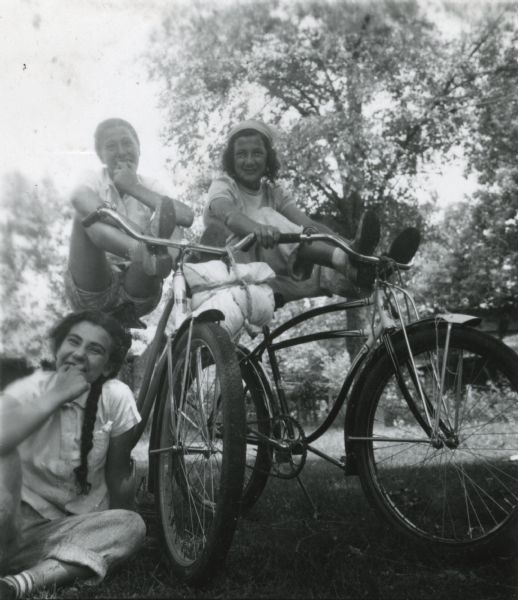 Two girls posing with their feet on the handlebars of their bikes, while one of the girls whistles with two fingers and a third girl (also whistling and mugging for the camera) sits next to them on the grass. The girls were part of a summer bike excursion program sponsored by Neighborhood House.