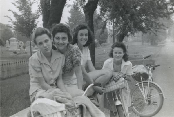 Four participants (Mary Lou Ciulla, Annie Gambino, Bessie Parisi, and Vita Valenza) of a bike hosteling trip sponsored by Neighborhood House, resting on a low wall and smiling for the camera.