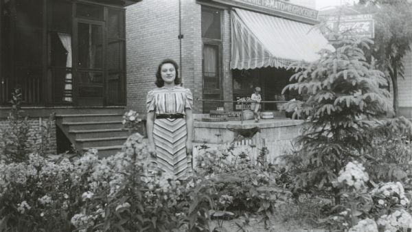 Image from the Garden Club record book kept by Neighborhood House, with the winner of the Best Garden competition in the Junior Girls category, Catherine LaBruzzo, standing in the garden of her house at 708 Milton Street. Next door is Amato Groceries, with a little boy and a dog outside the store.