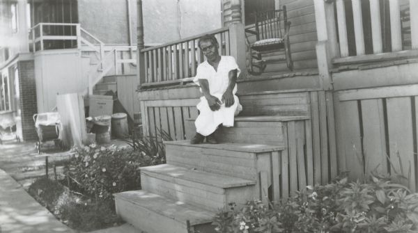Image from the Garden Club record book kept by Neighborhood House, with the winner in the Senior category garden competition, Mrs. Ora Smith, sitting on the front steps of her home at 212 S. Murray Street. Garden plots with flowers flank either side of the front steps to the house.