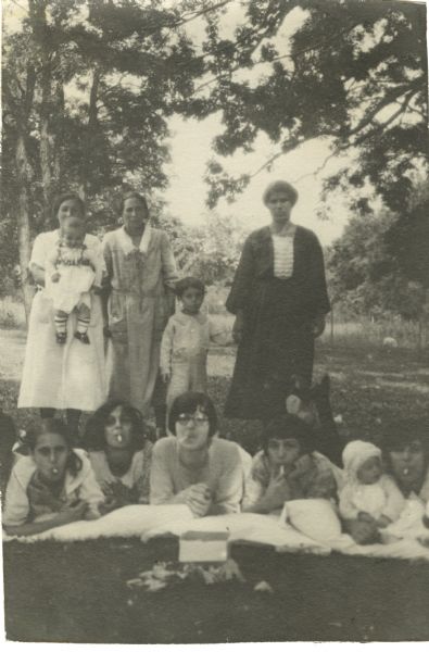 Mothers and small children standing and posing in Vilas Park, with other mothers and older children lying prone, propping themselves up with their elbows, resting on pillows, and pretending to smoke "cigarettes." This image is part of an album kept by Mary Lee Griggs, head of Parent Education and the Play School at Neighborhood House.