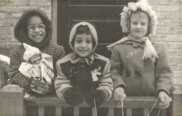 Three girls posing in winter jackets, hoods, and mittens, standing behind a railing and in front of the door to a building. One of the girls is holding up a doll. This image is part of an album kept by Mary Lee Griggs, head of Parent Education and the Play School at Neighborhood House.
