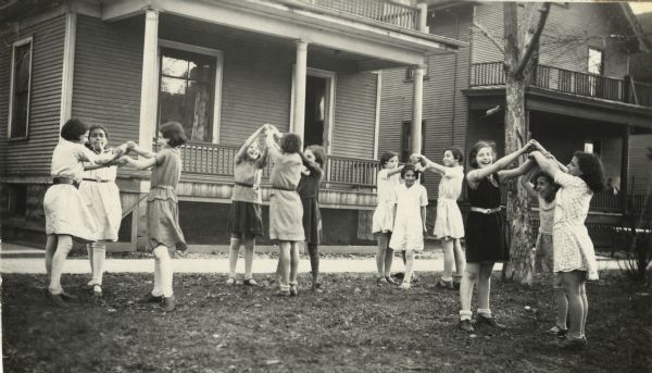 Four groups of girls in threes playing the game "London Bridge Is Falling Down," on the boulevard in front of a house. This image is part of an album kept by Mary Lee Griggs, head of Parent Education and the Play School at Neighborhood House.
