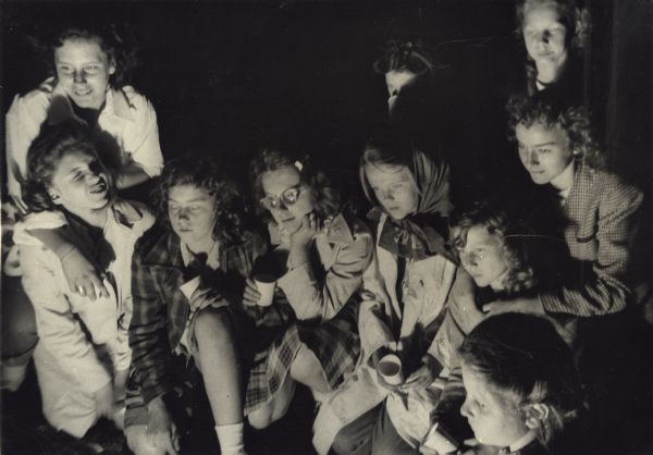 Group of girls in jackets, sitting or kneeling, perhaps illuminated by a campfire; some are holding cups. This image is from a hosteling scrapbook kept by Mary Lee Griggs, head of Parent Education and the Play School at Neighborhood House. The caption on the page reads, "The Gang — At our Scavenger Hunt and Apple Cider Party — Brittingham Park."