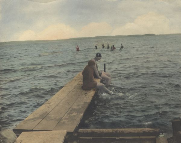 Hand-colored image of two swimmers sitting on a pier with their legs in the water at a camp in West Point sponsored by Neighborhood House. Several other swimmers are standing further out in the lake.