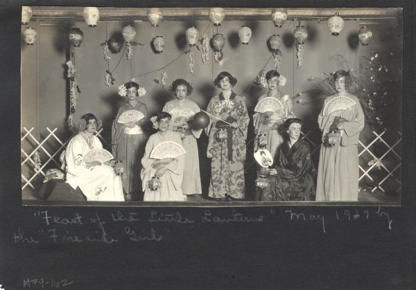 Page from a scrapbook kept by Neighborhood House, with cast members of "Feast of the Little Lanterns," a production staged by the Fireside Girls club. The girls are wearing kimonos and flowers in their hair, and holding fans and small lanterns, beneath a stage set hung with many larger lanterns.