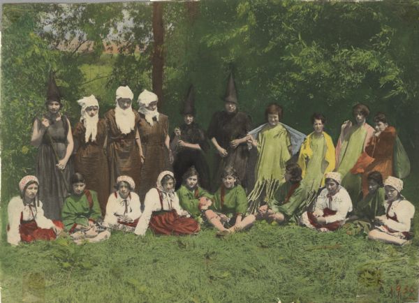 Hand-colored image from an scrapbook kept by Neighborhood House, with cast members of a production with nymphs, witches, village boys and girls, and a peddler (Katie Caramella?, standing, far right), posing outdoors.