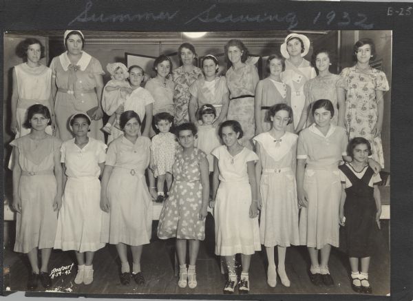 Group of girls in a summer sewing group at Neighborhood House, posing in their finery with Gay Braxton (back row, center, under ceiling lamp), head resident of the settlement house.