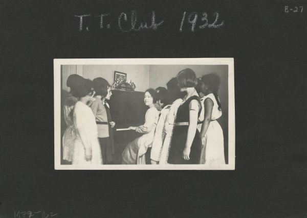 Page from an album kept by Neighborhood House, with T.T. Club members gathered around a piano while Mary Lee Griggs, director of the Play School, plays accompaniment.