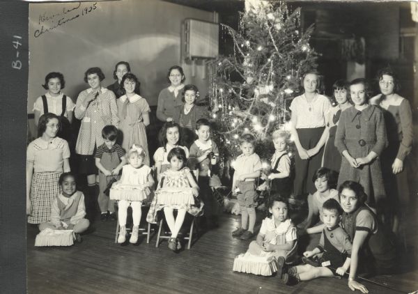 Page from a scrapbook kept by Neighborhood House, with Kiwalan Camp Fire girls with preschoolers, posing at a Christmas celebration near a tree decorated with tinsel, garlands, and lights. The smaller children in the front rows are holding small toys: doll beds for the girls and stick toys for the boys.