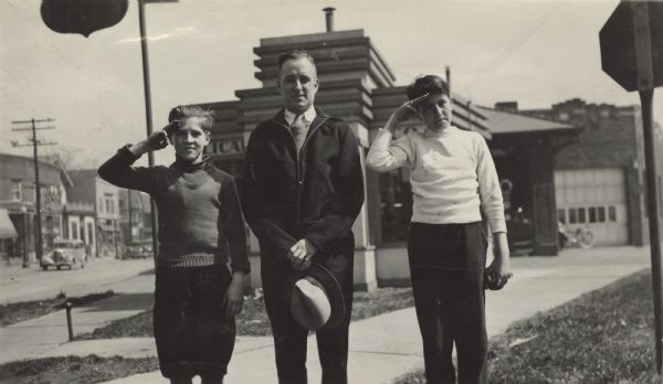 Image from a scrapbook kept by Neighborhood House, with a three-quarter view of a man and two boy scouts on either side of him who are standing at attention and saluting. The buildings behind them are the Licari Tire Company office and garage, at 767 W. Washington Avenue, across the street from the settlement house. A Phillips sign is hanging from a post in the tire company's yard.