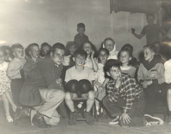 A boy with boxing gloves sitting and awaiting his turn in the ring, with a man and another boy crouching on either side of him and other children behind them sitting along a wall. Family Nights were held the last Fridays of the month at the settlement house, with activities for children and their families.