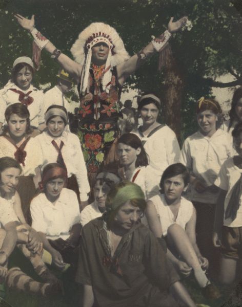 Hand-colored group portrait of Camp Fire girls and a man dressed in Ojibwa-style floral embroidered/beaded clothing and a Plains Indian feathered headdress and standing with upraised arms, taken at Camp The Dells. Other adults are visible in the background.