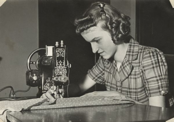 Image from a scrapbook kept by Neighborhood House, with Lucy Martinelli stitching a seam using an elaborately decorated electric sewing machine. Sewing classes were a frequent offering at the settlement house.