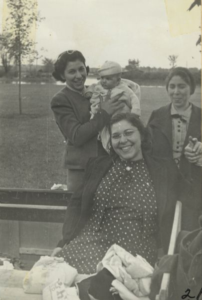 Image from a scrapbook kept by Neighborhood House, with Jennie G., Rose Caruso, and Mrs. Urso at a picnic. One of the women is sitting on a park bench, while one of the two women standing behind her is holding a baby.