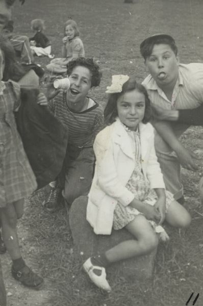 Image from a scrapbook kept by Neighborhood House, with Mary Vultaggio sitting on a rock with two boys behind her who are clowning for the camera (Frank Romano is sticking out his tongue, and other children around them on the grass.