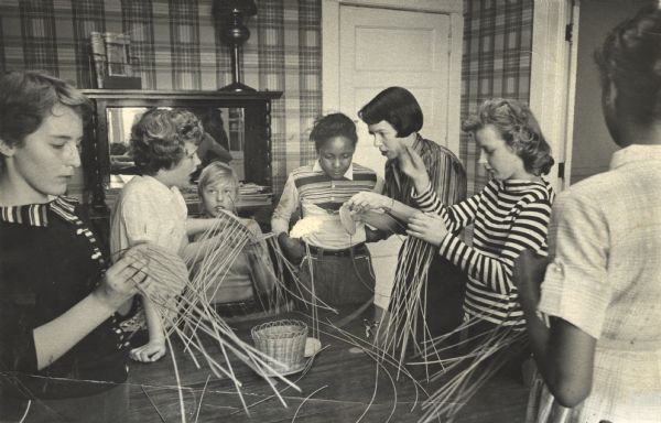 Group of girls making baskets, as an older girl or woman demonstrates a technique. Neighborhood House sponsored many groups and activities, such as basketry, needle arts, and woodwork, for children.
