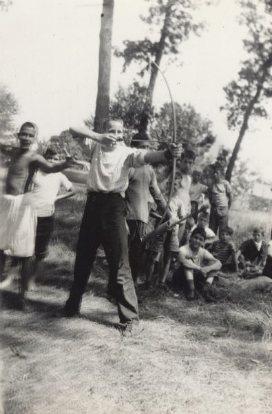 Image from a scrapbook kept by Neighborhood House, with Pat McCann, the last shooter in an archery tournament held at a boys summer camp, drawing the bow and poised to shoot.