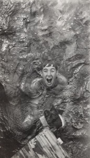 Image from a scrapbook kept by Neighborhood House of a boys summer camp, with an image of Skip Sul?(obscured letters) participating in a game involving taking off the pajamas he is wearing while in the water, throwing them to a team member who dons them, swims a distance, returns to shore, and then removes the wet garments.