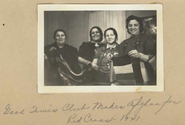 Image from a scrapbook kept by Neighborhood House, with women from the Good Times Club holding up an afghan they made for the Red Cross. A group of women, often with sons in military service, met weekly at the settlement house to sew and knit items for the relief organization.