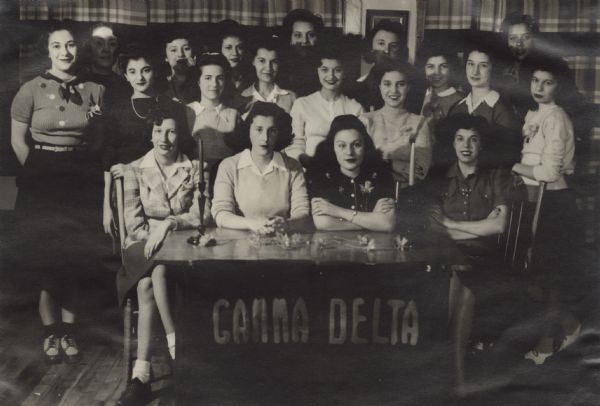 Group portrait of members of a club for teenage girls at Neighborhood House, with four girls seated at a table with a banner that reads "Gamma Delta," and two rows of girls standing behind them.