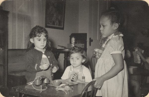 Image from a scrapbook kept by Neighborhood House, with three girls standing and sitting at a table with a play telephone, a kitchen beater, and play dishes. Two of the girls are wearing necklaces made of large wooden beads. Other children are stacking blocks at another table in the background.