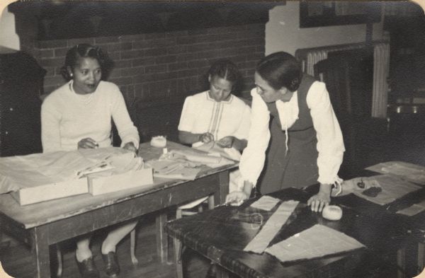 Image from a scrapbook kept by Neighborhood House, with three women working on sewing pattern pieces: two women are sitting at a table with a box of pattern sheets, with one woman sewing; the third woman stands at another table, poised to cut out pattern pieces pinned to cloth.