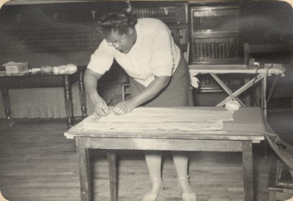 Image from a scrapbook kept by Neighborhood House, with a woman standing over a table, pinning a sewing pattern piece to cloth. An ironing board and bookcases can be seen in the background.