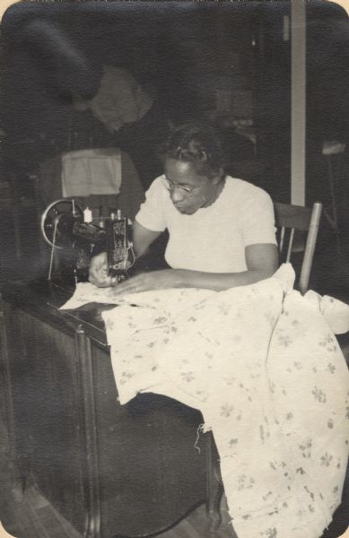 Image from a scrapbook kept by Neighborhood House, with a woman using a sewing machine to sew a large piece of cloth patterned with four-leaf clovers. Another woman can be seen in the background, pinning pattern pieces to cloth.